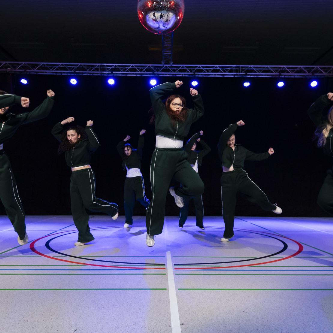 Sechs T&auml;nzerinnen in sportlicher Kleidung f&uuml;hren synchron eine Choreografie auf einer beleuchteten Tanzfl&auml;che aus.