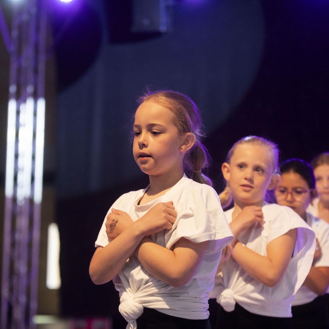 Sechs Kinder in wei&szlig;en T-Shirts f&uuml;hren einen Tanz auf einer B&uuml;hne mit buntem Licht auf.