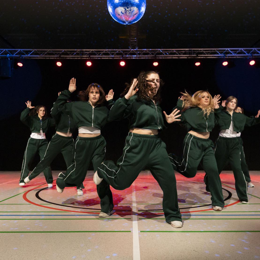 Sechsk&ouml;pfige Tanzgruppe in gr&uuml;nen Trainingsanz&uuml;gen tanzt energetisch auf einer B&uuml;hnenfl&auml;che mit Discokugel und Lichtern.