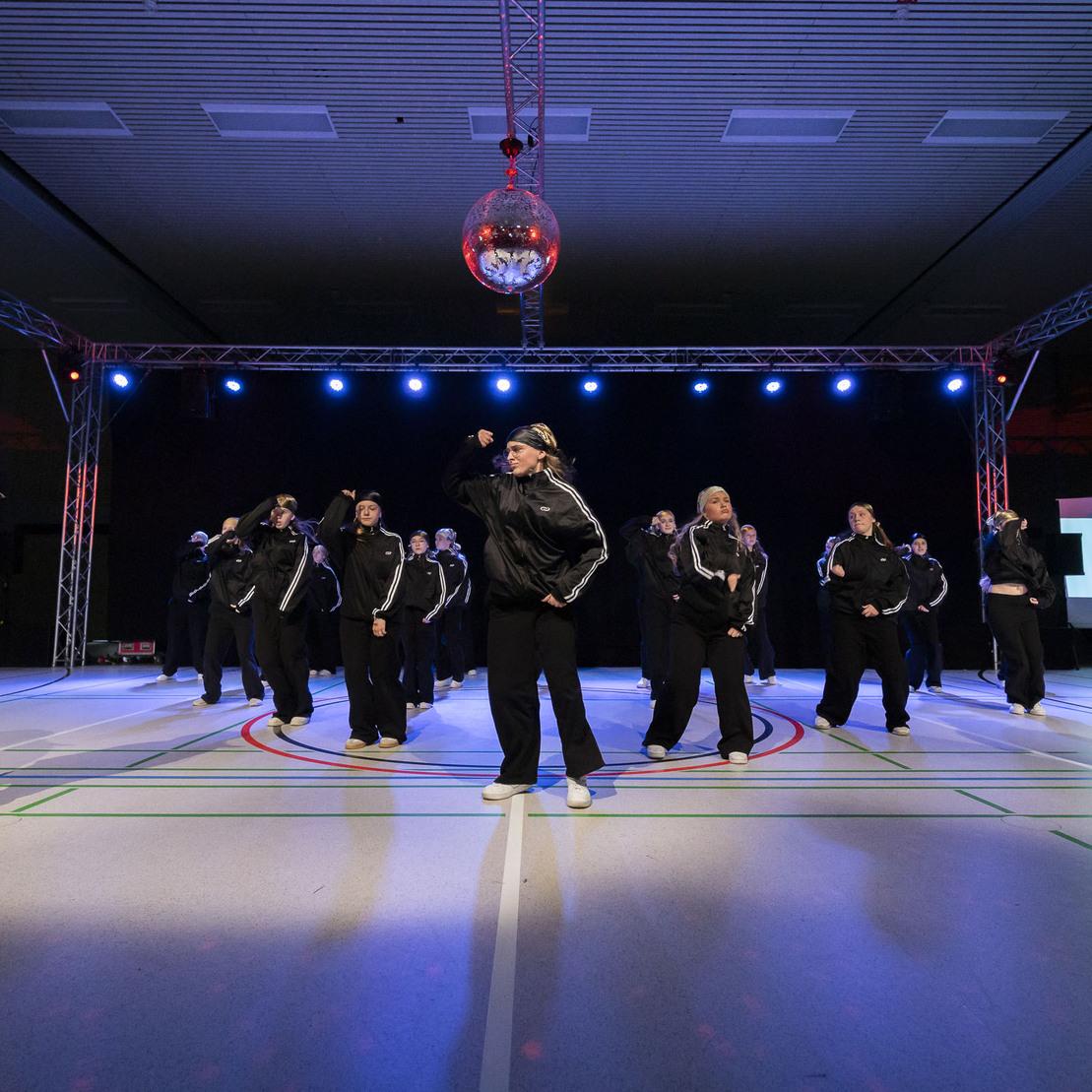 Tanzgruppe in schwarzen Outfits f&uuml;hrt eine choreografierte Auff&uuml;hrung unter bunten Lichtern auf einer B&uuml;hne aus.