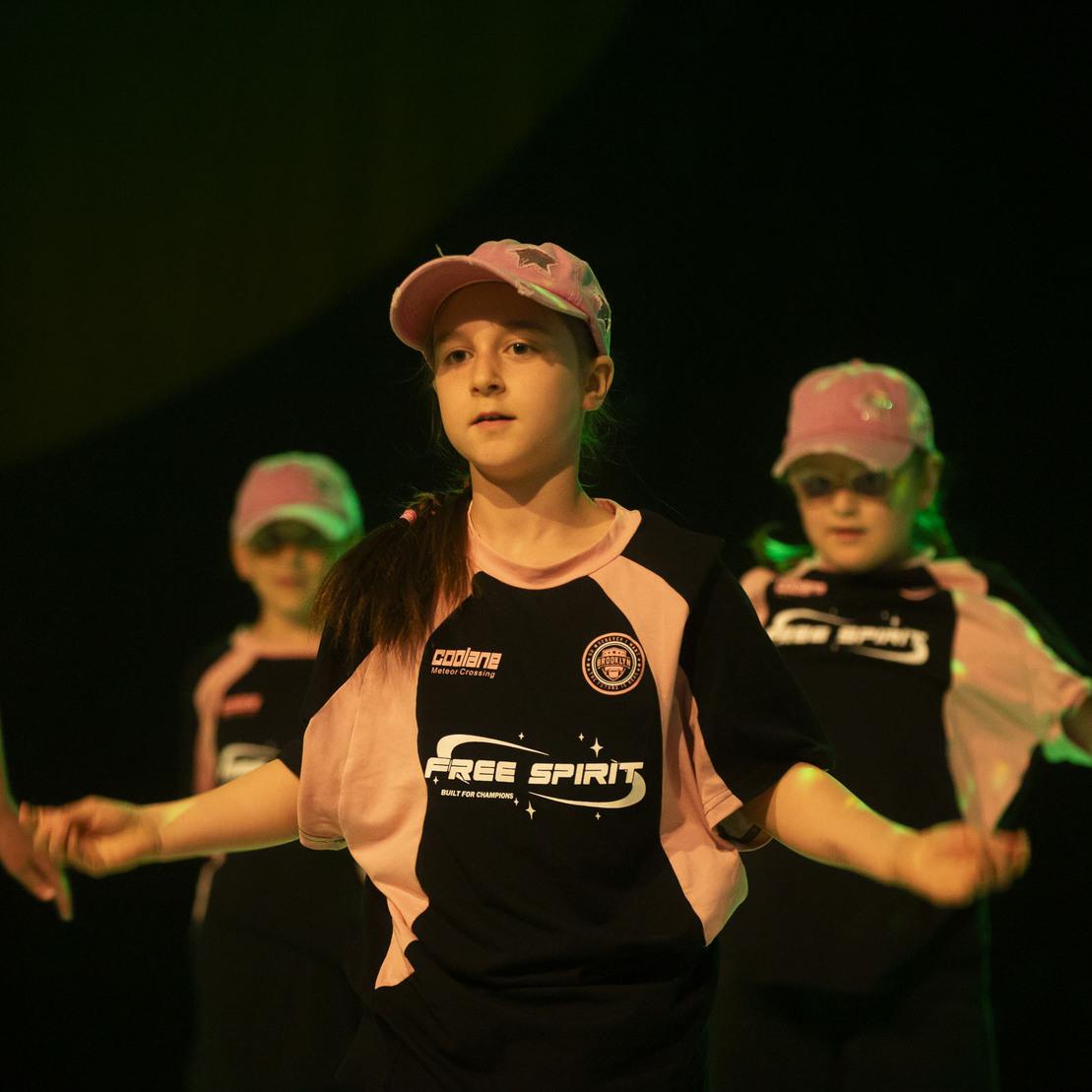 Tanzende Kinder in einheitlicher Kleidung und M&uuml;tzen f&uuml;hren eine Choreografie auf einer B&uuml;hne mit schwachem Licht aus.