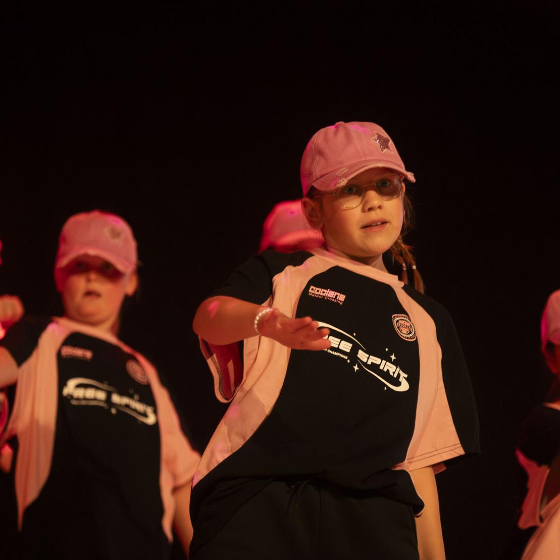 Gruppe von M&auml;dchen in schwarzen Tanz-Shirts und rosa Kappen f&uuml;hrt eine Choreografie auf einer B&uuml;hne vor.