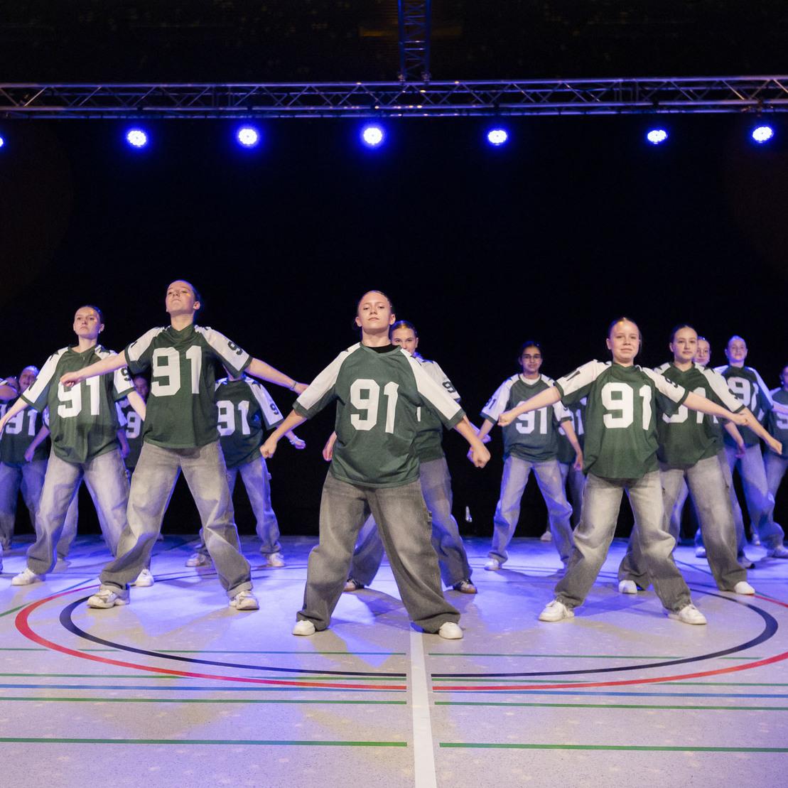 Tanzgruppe in gr&uuml;nen Trikots mit der Zahl 91 f&uuml;hrt eine synchronisierte Choreografie auf einer B&uuml;hnenfl&auml;che aus.