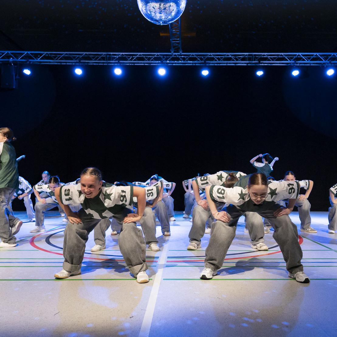 Tanzgruppe f&uuml;hrt eine choreografierte Routine auf einer B&uuml;hne mit blauem Licht und Spiegelkugel auf.
