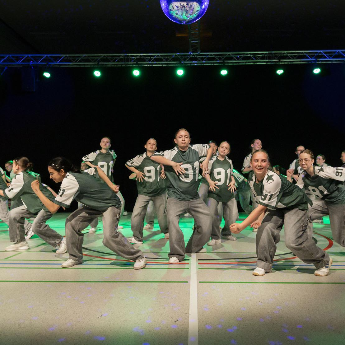Tanzgruppe auf einer B&uuml;hne in Einheitskleidung, die synchron eine Choreografie mit lebhaften Bewegungen auff&uuml;hrt.