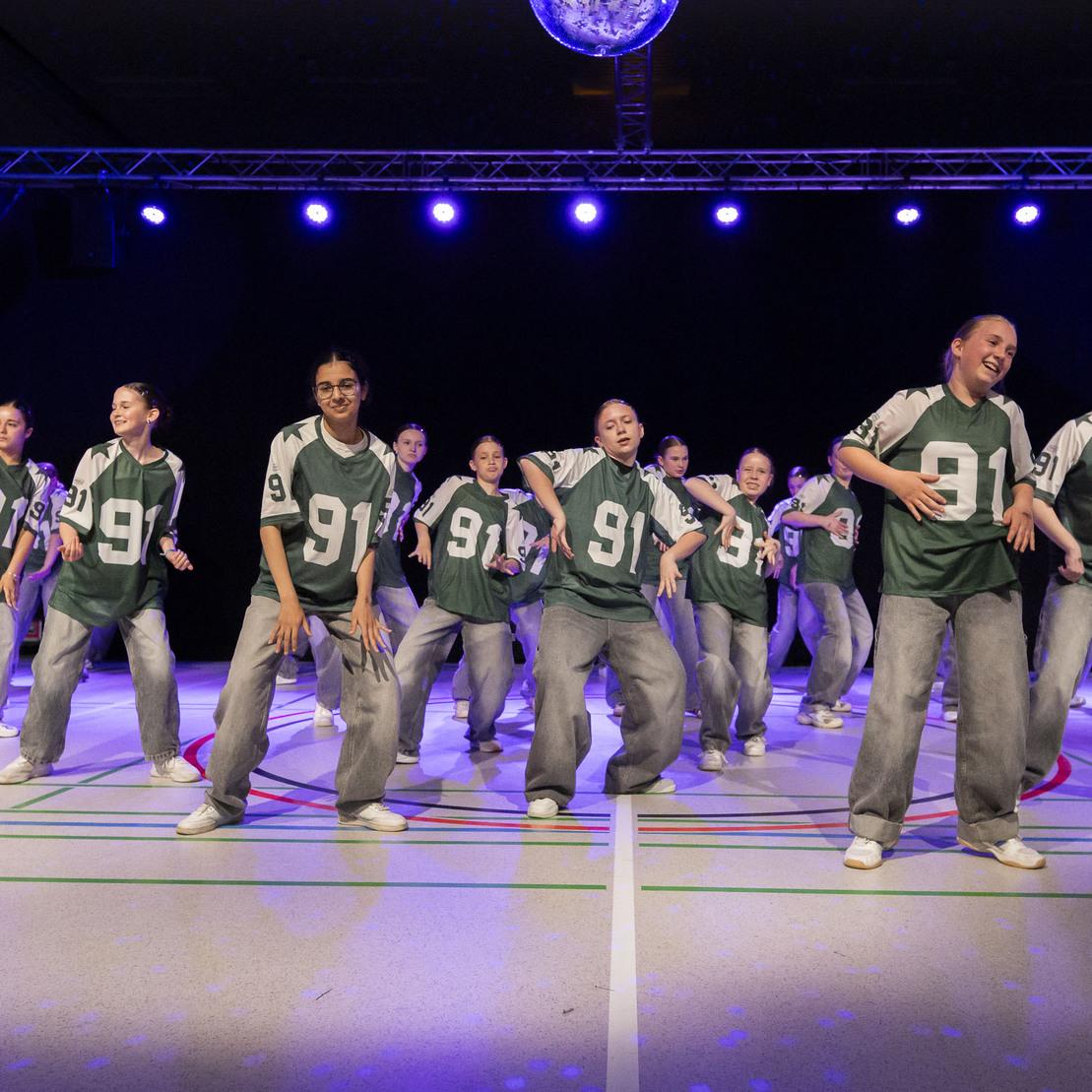 Tanzgruppe in gr&uuml;nen Trikots mit der Nummer 91, die auf einer B&uuml;hne unter buntem Licht performt.