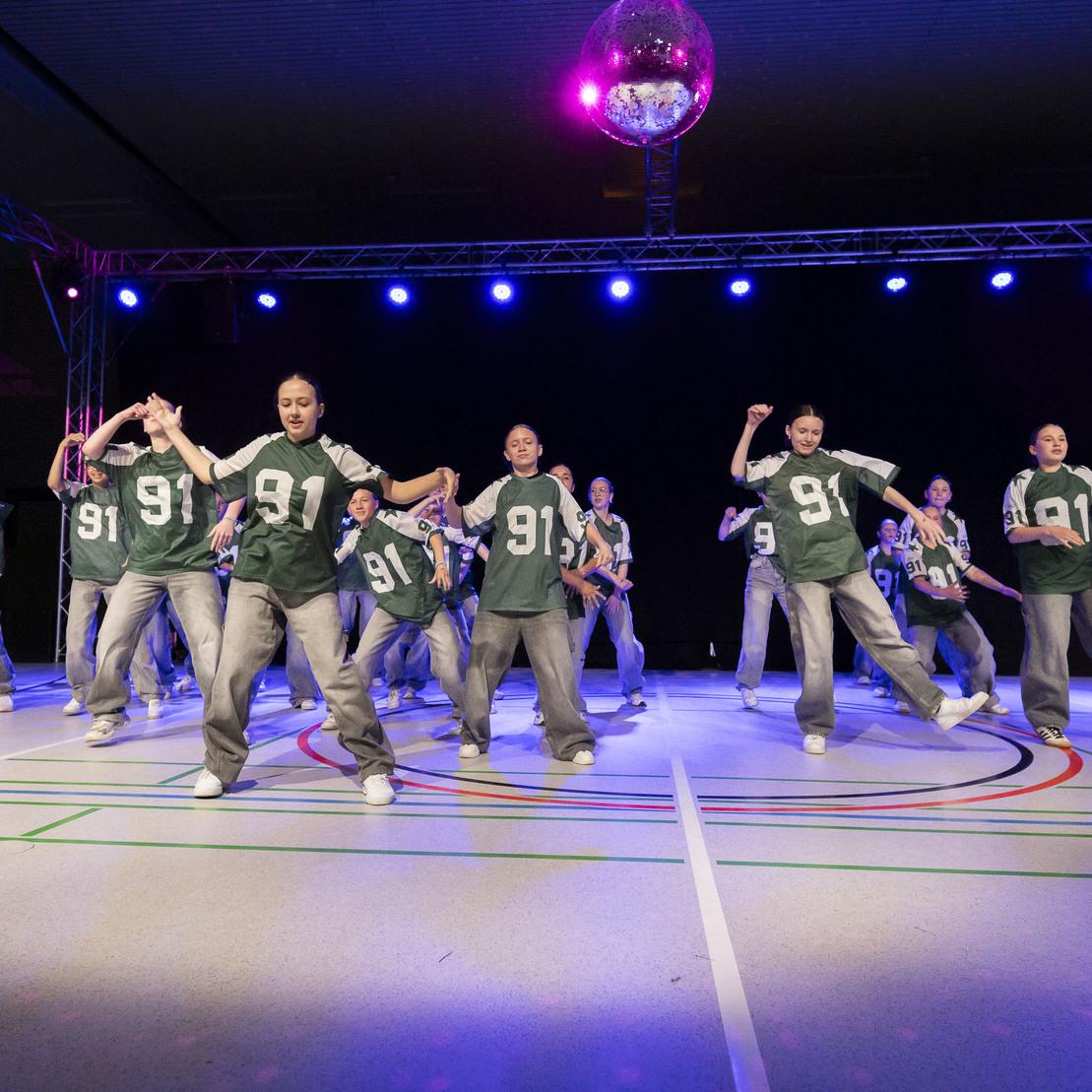 Tanzgruppe in einheitlichen gr&uuml;nen Shirts f&uuml;hrt einen energetischen Auftritt unter bunten Lichtern auf einer Sportfl&auml;che durch.