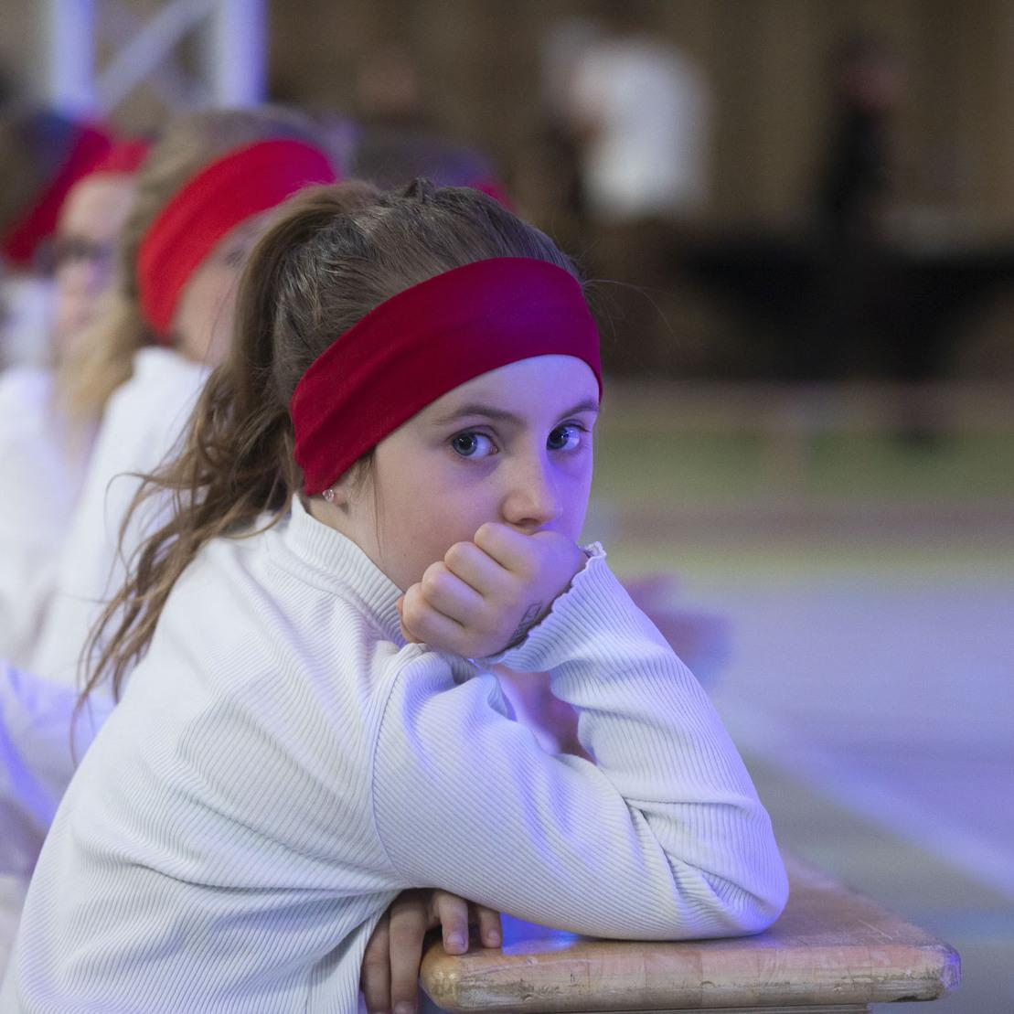 M&auml;dchen mit rotem Stirnband sitzt am Tisch und schaut nachdenklich in die Kamera, im Hintergrund weitere Kinder.