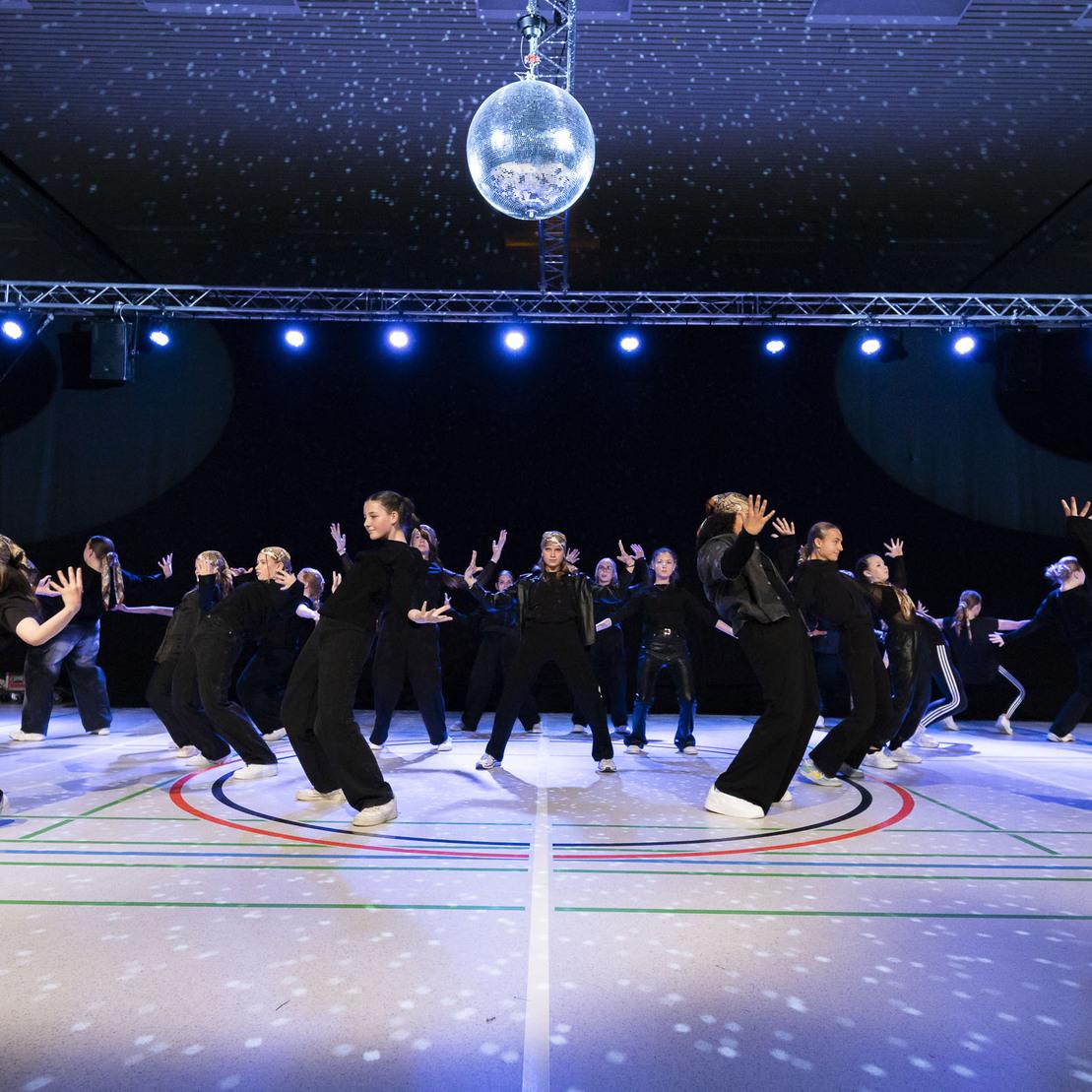 Tanzgruppe in eleganter Formation auf einer B&uuml;hne mit Spiegelkugel und stimmungsvollem Licht in einer Sporthalle.
