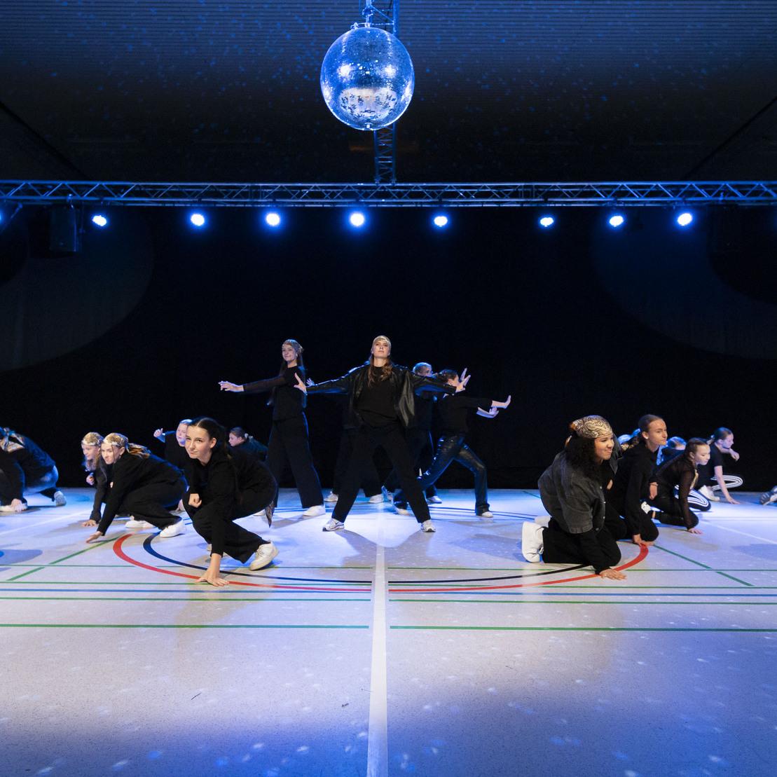 Tanzauff&uuml;hrung in einer Halle mit mehreren T&auml;nzern in Schwarz unter bunten Lichtern und einer Discokugel.