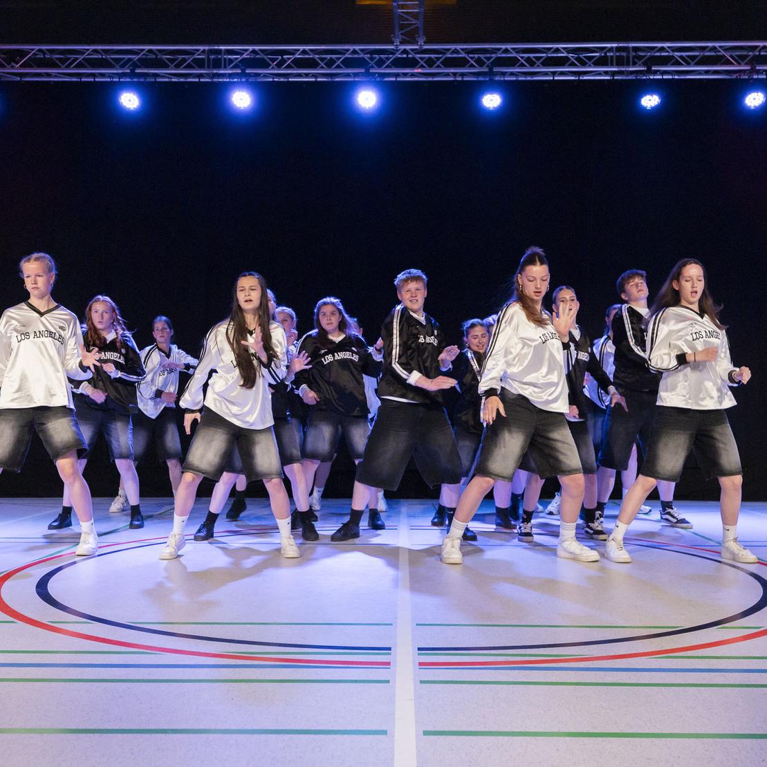 Tanzgruppe in sportlichen Outfits f&uuml;hrt eine Choreografie auf einer beleuchteten B&uuml;hne mit Farbakzenten auf.