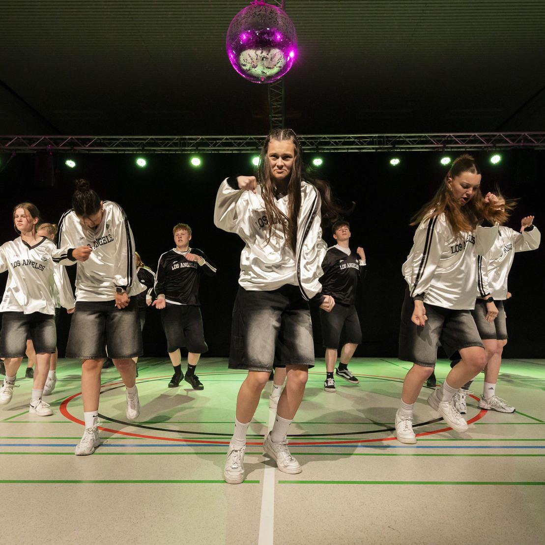 Gruppe von T&auml;nzern in silbernen Outfits f&uuml;hrt eine Choreografie unter bunten Lichtern auf einer Sporthallenfl&auml;che vor.