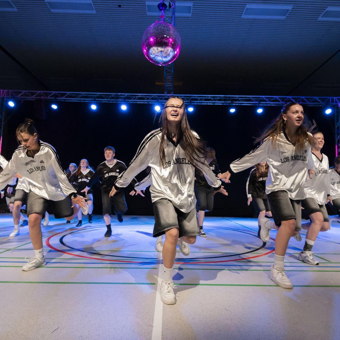 Tanzgruppe in Silber-Shirts f&uuml;hrt synchronen Tanz auf einer B&uuml;hne mit Disco-Kugel und bunten Lichtern auf.