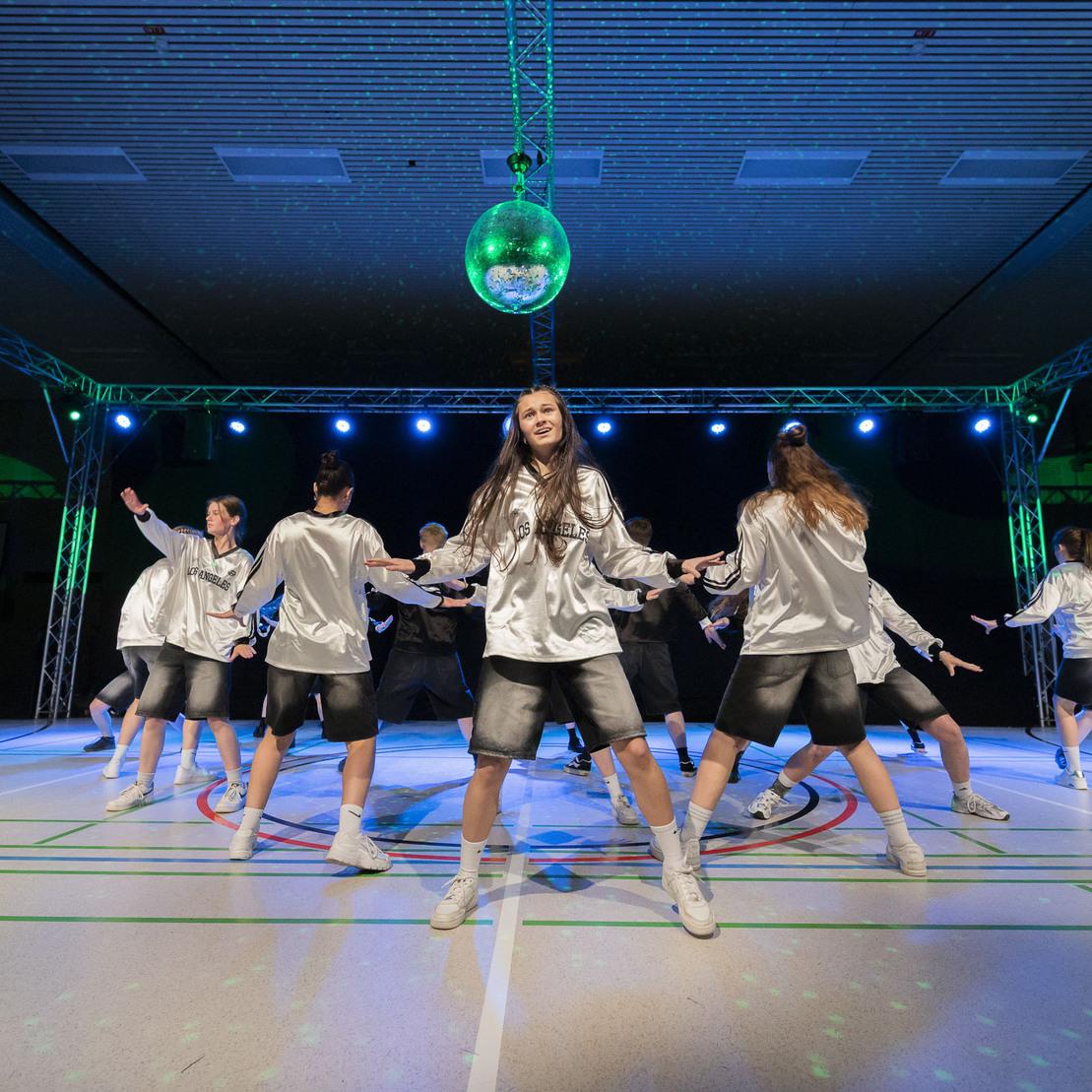 T&auml;nzerinnen in silbernen Oberteilen und Shorts f&uuml;hren eine Choreografie auf einer beleuchteten Sportfl&auml;che auf.