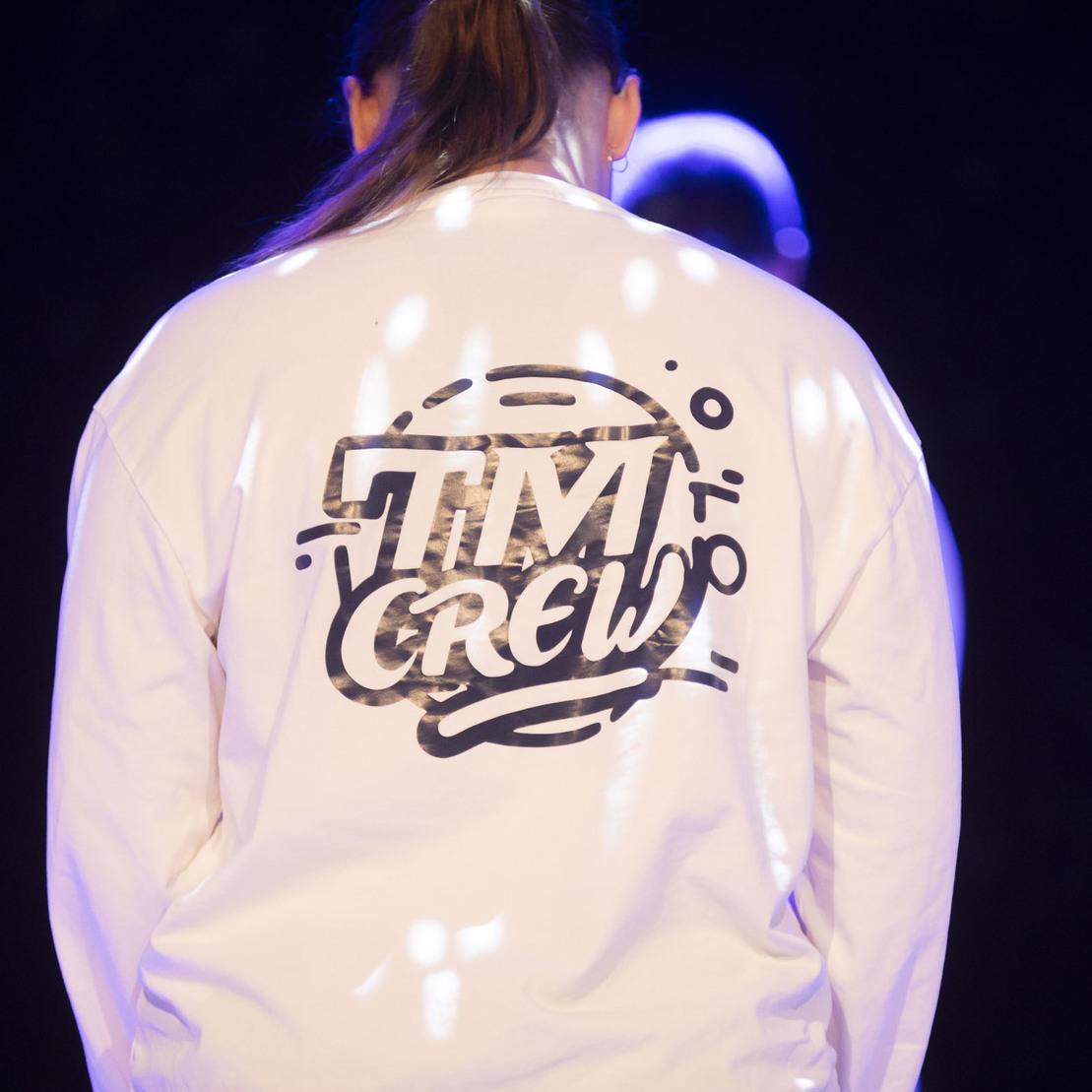 Nahaufnahme eines Oberteils mit dem Schriftzug &bdquo;TIM CREW&ldquo; auf dem R&uuml;cken, beleuchtet von bunten Lichtern.