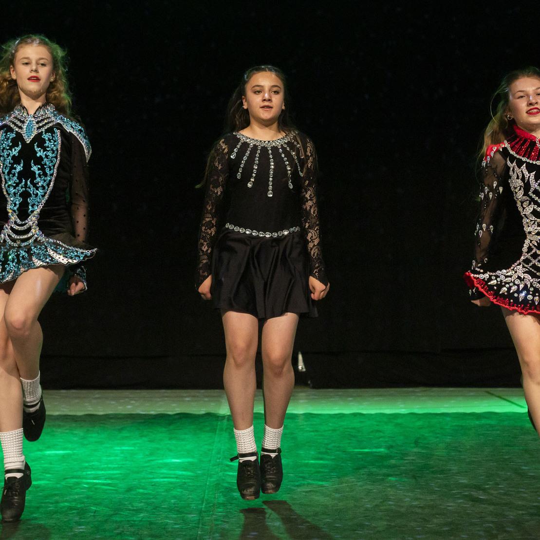 Drei T&auml;nzerinnen in traditioneller irischer Tanzkleidung f&uuml;hren synchron einen Tanz auf einer beleuchteten B&uuml;hne vor.