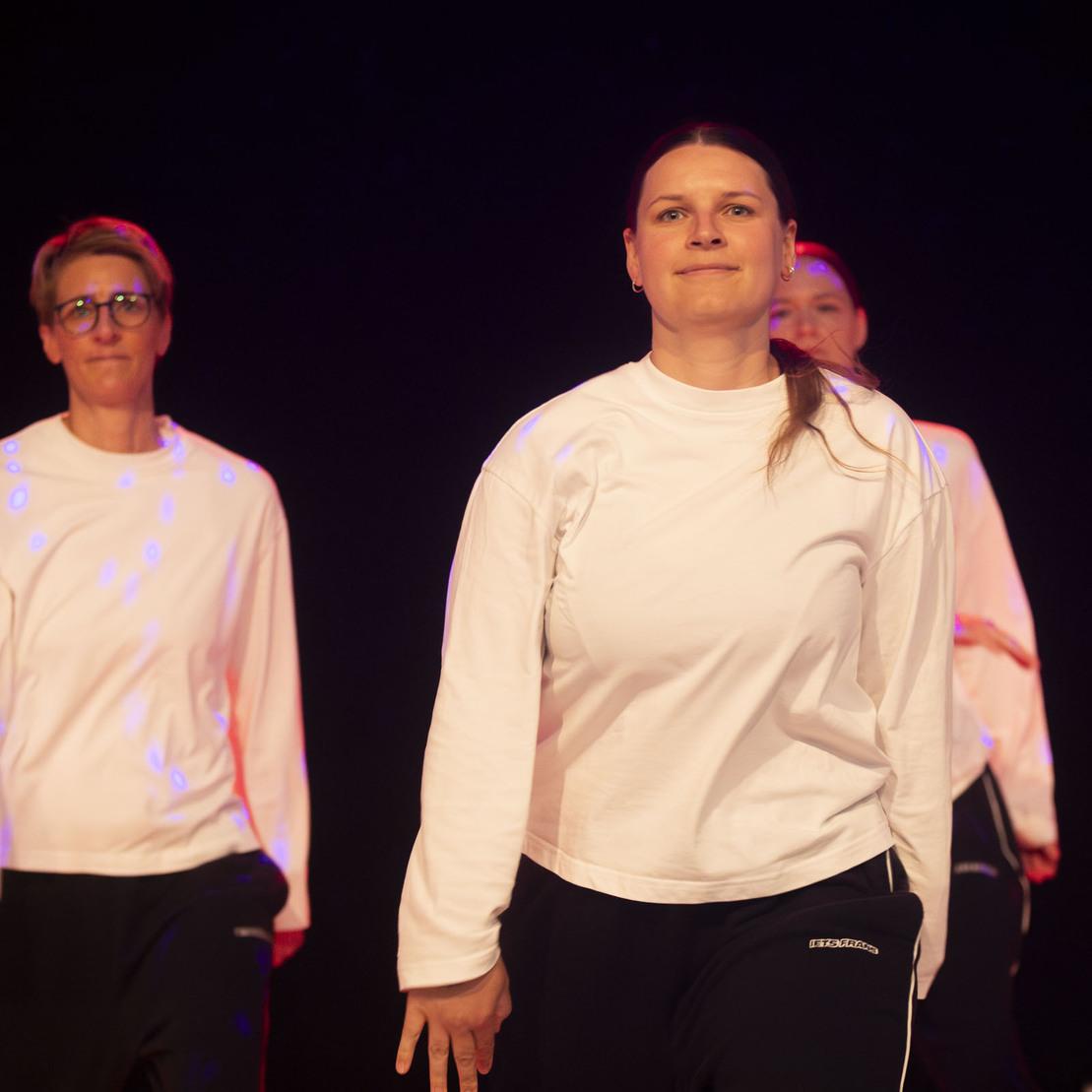 Drei Frauen in wei&szlig;en Sweatshirts und Dunkelhosen tanzen auf einer B&uuml;hne mit roten und blauen Lichtakzenten.