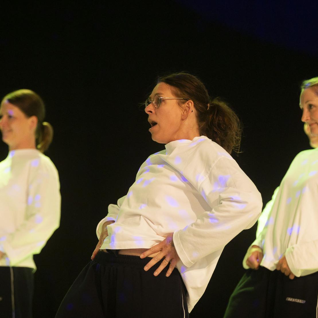 Drei T&auml;nzerinnen in wei&szlig;en Sweatshirts f&uuml;hren eine Choreografie vor einem dunklen Hintergrund auf.