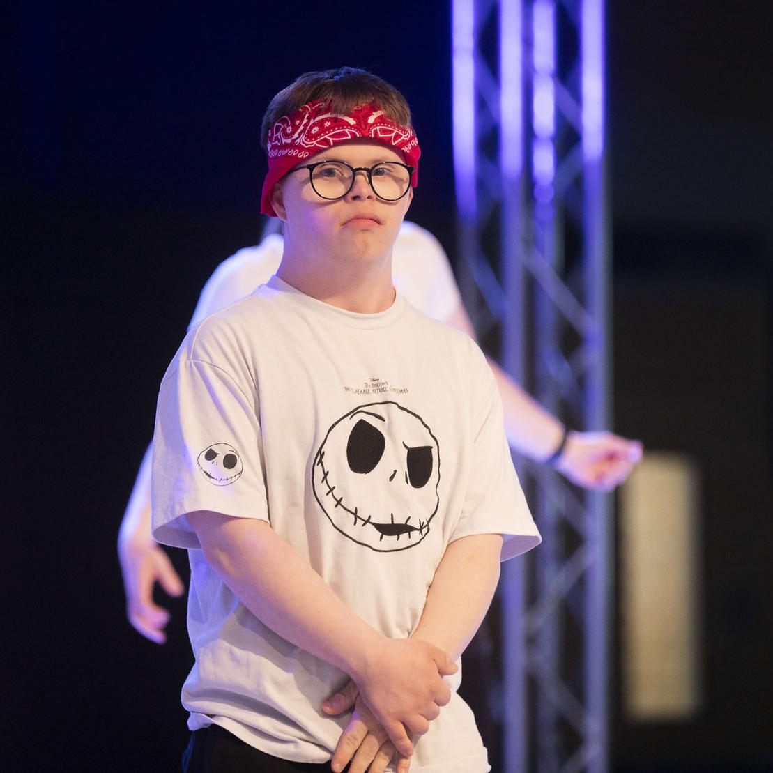 T&auml;nzer mit Brille und rotem Bandana in einem wei&szlig;en Shirt mit Motiv steht auf einer B&uuml;hne, umgeben von anderen T&auml;nzern.