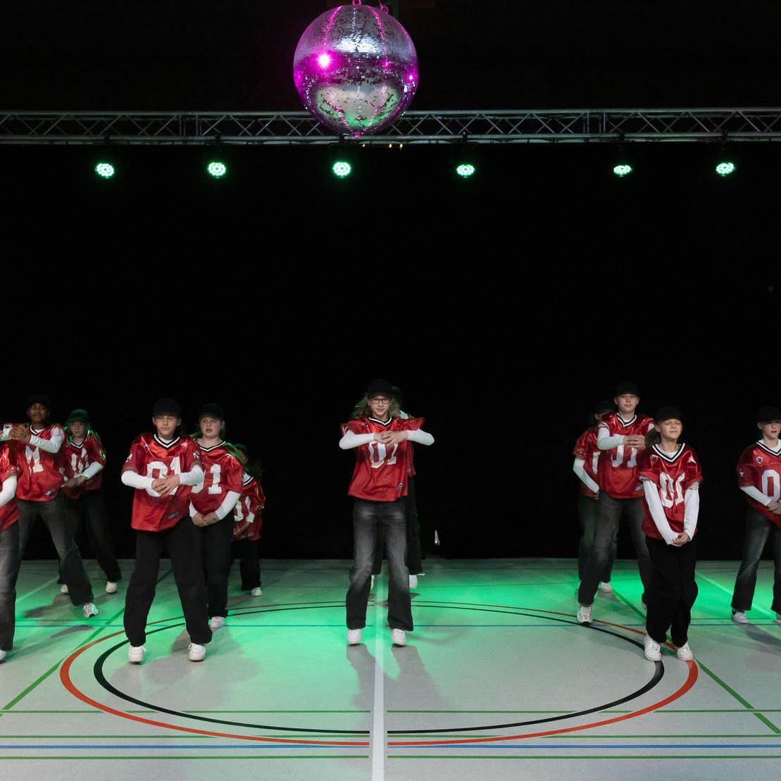 Gruppe von T&auml;nzern in roten Trikots f&uuml;hrt eine Choreografie unter einem Diskokugel-Licht auf einer beleuchteten Tanzfl&auml;che auf.
