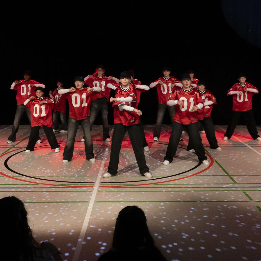 Tanzgruppe in roten Shirts f&uuml;hrt eine Choreografie auf einer beleuchteten B&uuml;hne mit Sportboden vor.
