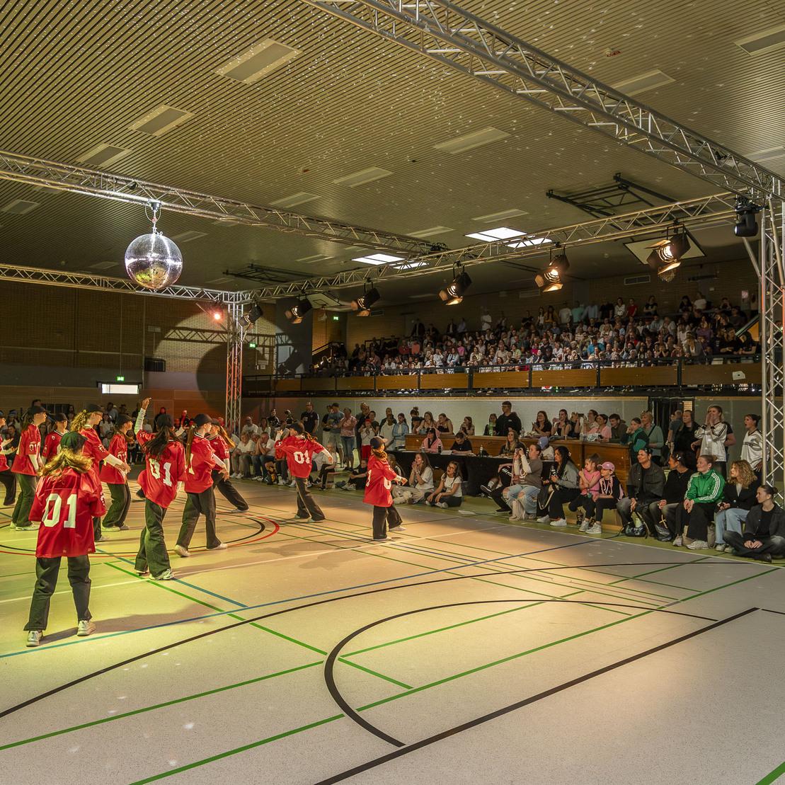 Tanzgruppe in roten Shirts f&uuml;hrt eine Choreografie in einer Sporthalle vor Publikum auf.