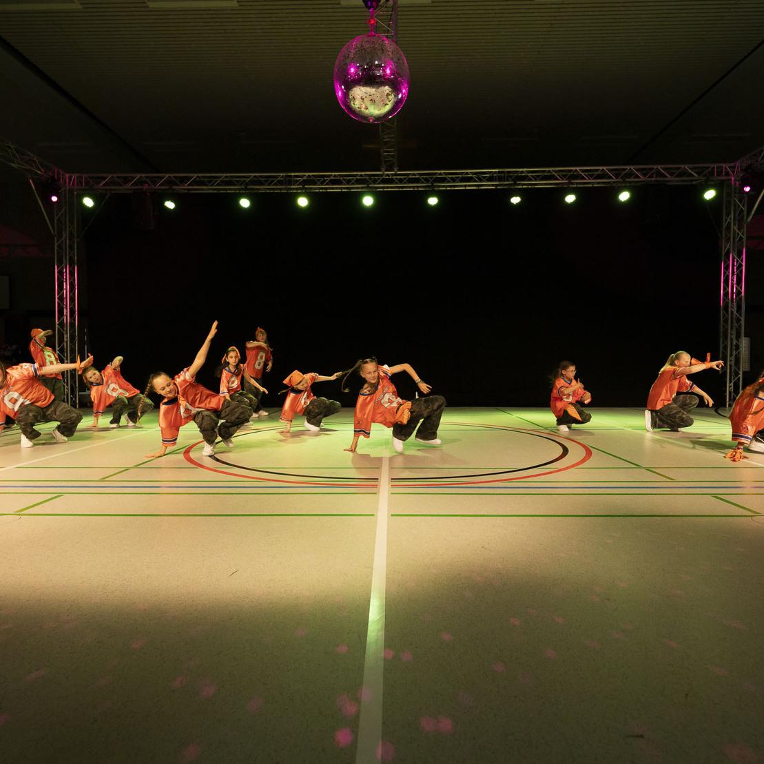 Tanzgruppe in roten Outfits f&uuml;hrt eine Performance auf einer beleuchteten B&uuml;hne mit Discokugel und gr&uuml;ner Fl&auml;che auf.