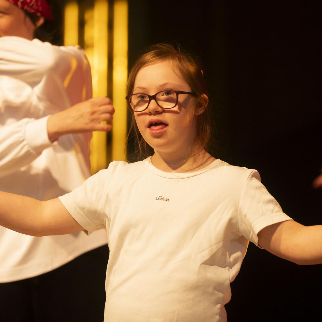 M&auml;dchen mit Down-Syndrom tanzt auf der B&uuml;hne, umgeben von anderen T&auml;nzern in wei&szlig;en T-Shirts.