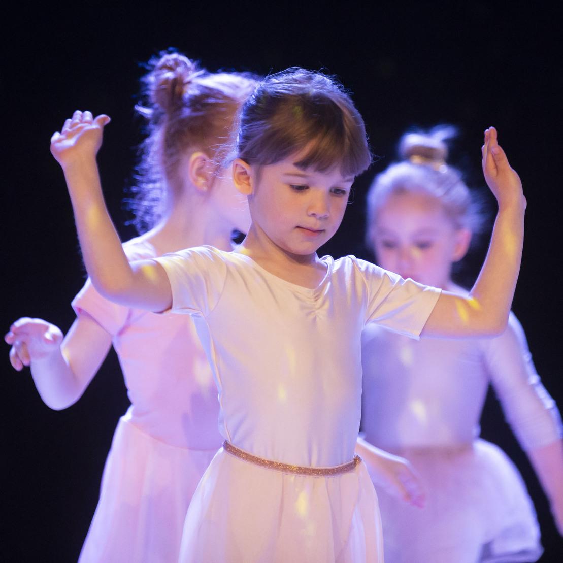 M&auml;dchen in Ballettkleidung tanzt im Vordergrund, w&auml;hrend zwei weitere T&auml;nzerinnen verschwommen im Hintergrund zu sehen sind.