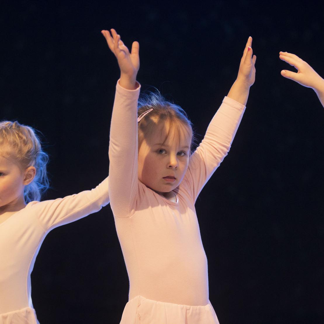 Drei M&auml;dchen in rosa Ballettanz&uuml;gen mit erhobenen Armen auf einer B&uuml;hne in dunkler Umgebung.