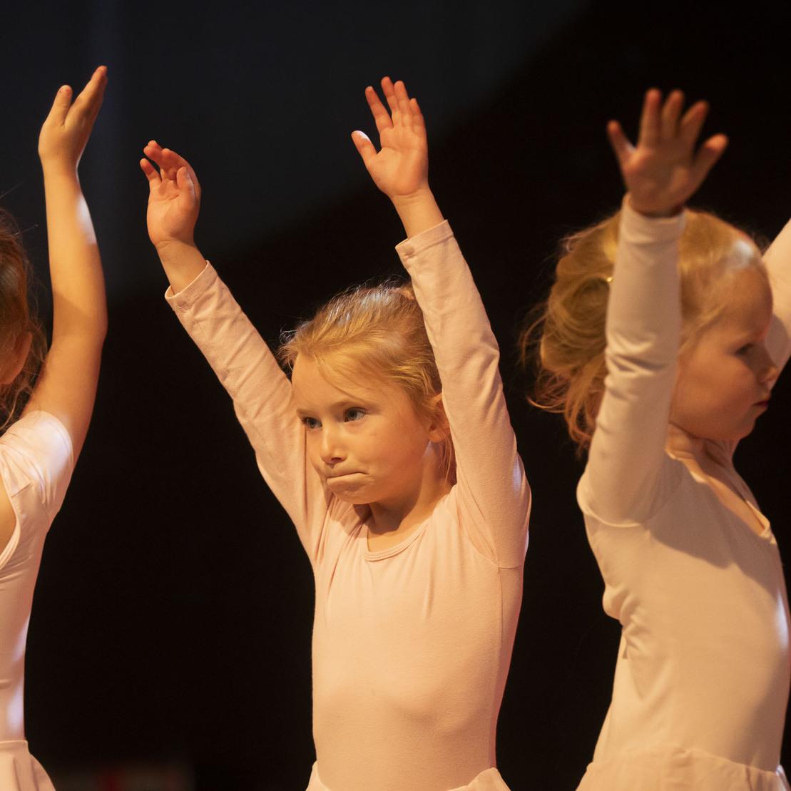 Drei junge Ballettt&auml;nzerinnen in rosa Kost&uuml;men heben die Arme w&auml;hrend eines Auftritts auf einer B&uuml;hne.
