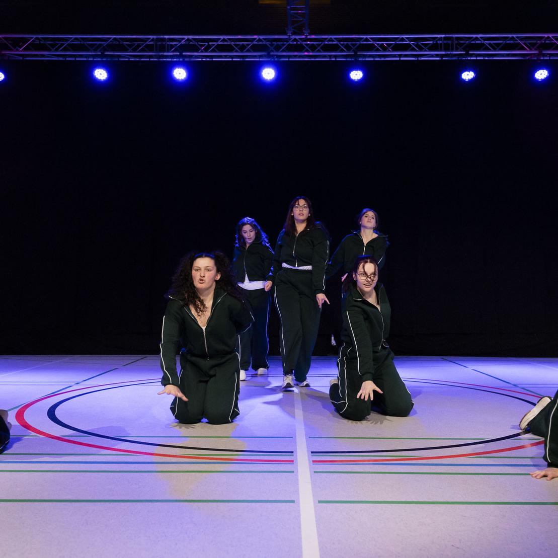 Sechs T&auml;nzerinnen in schwarzen Trainingsanz&uuml;gen f&uuml;hren eine Choreografie auf einem sportlichen Boden mit bunten Linien auf.