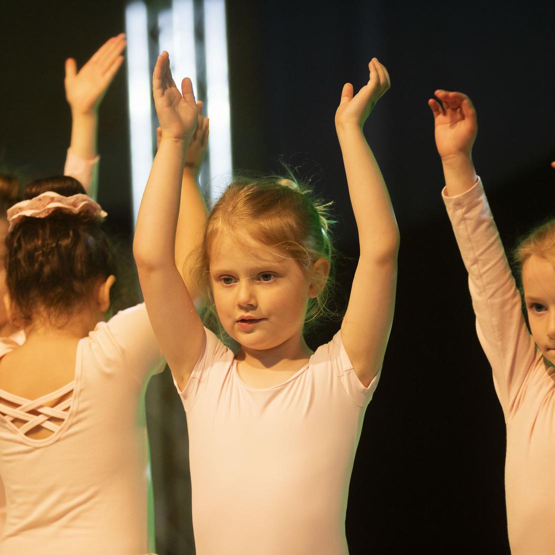 Drei kleine Ballett&auml;nzerinnen heben die Arme in einer Auff&uuml;hrung, tragen rosa Trikots und stehen auf einer B&uuml;hne.