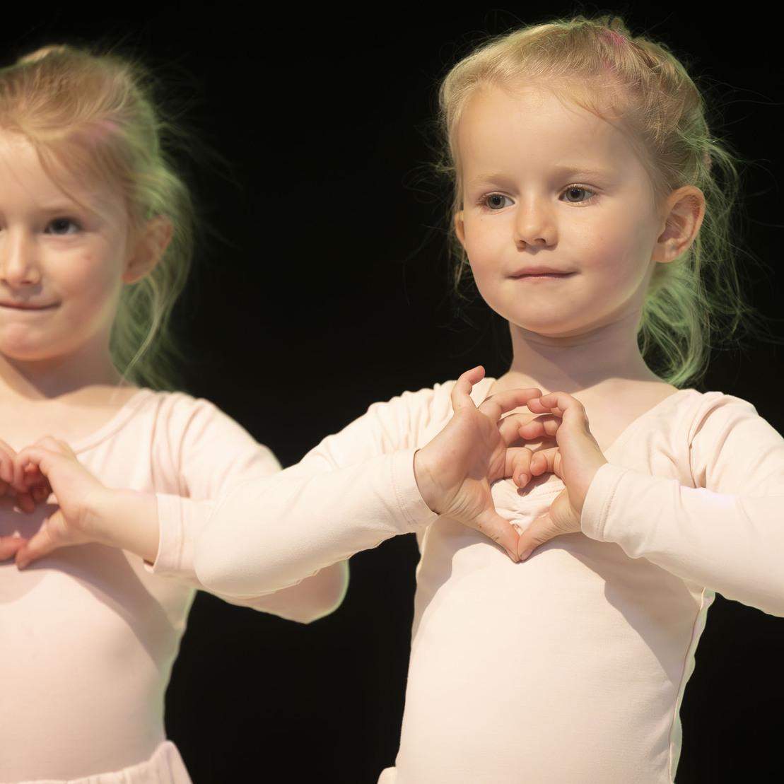 Zwei kleine T&auml;nzerinnen in rosa Ballettanz&uuml;gen formen mit ihren H&auml;nden Herzsymbole auf einer B&uuml;hne.