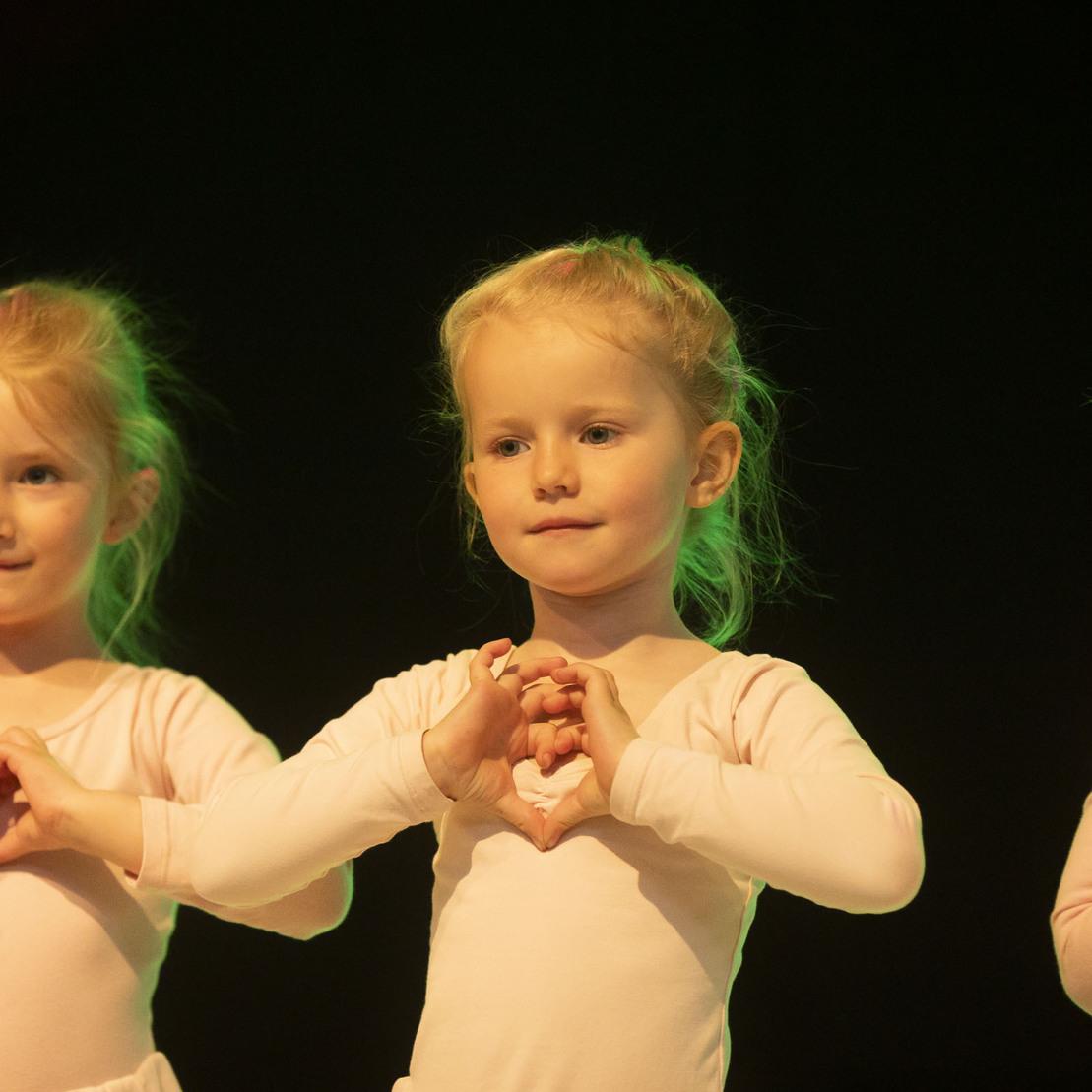 Drei kleine M&auml;dchen in rosa Ballettanz&uuml;gen formen mit ihren H&auml;nden Herzen auf einer B&uuml;hne.