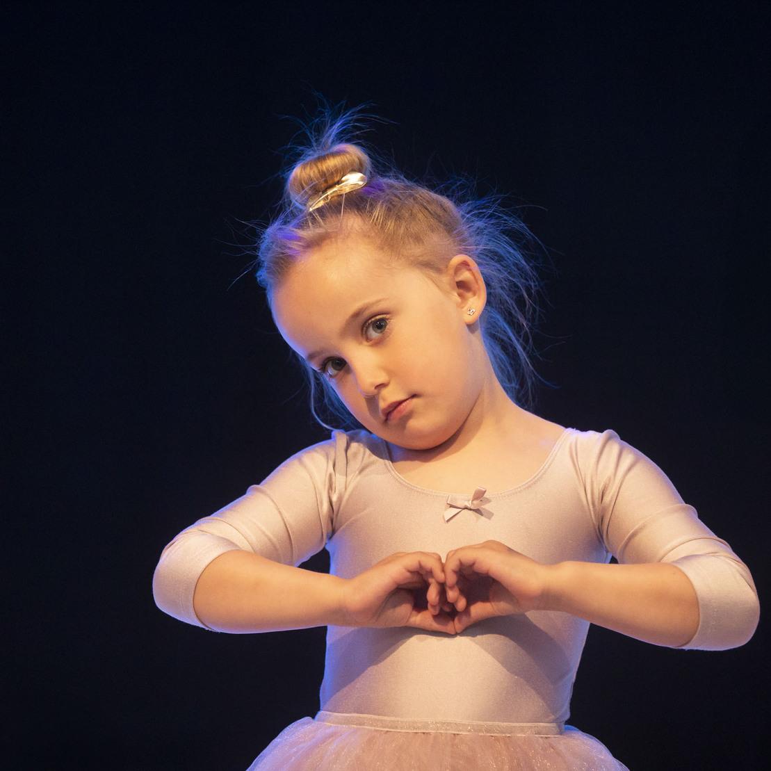 M&auml;dchen in Ballettkost&uuml;m formt mit den H&auml;nden ein Herz, sanft beleuchtet von einem dunklen Hintergrund.