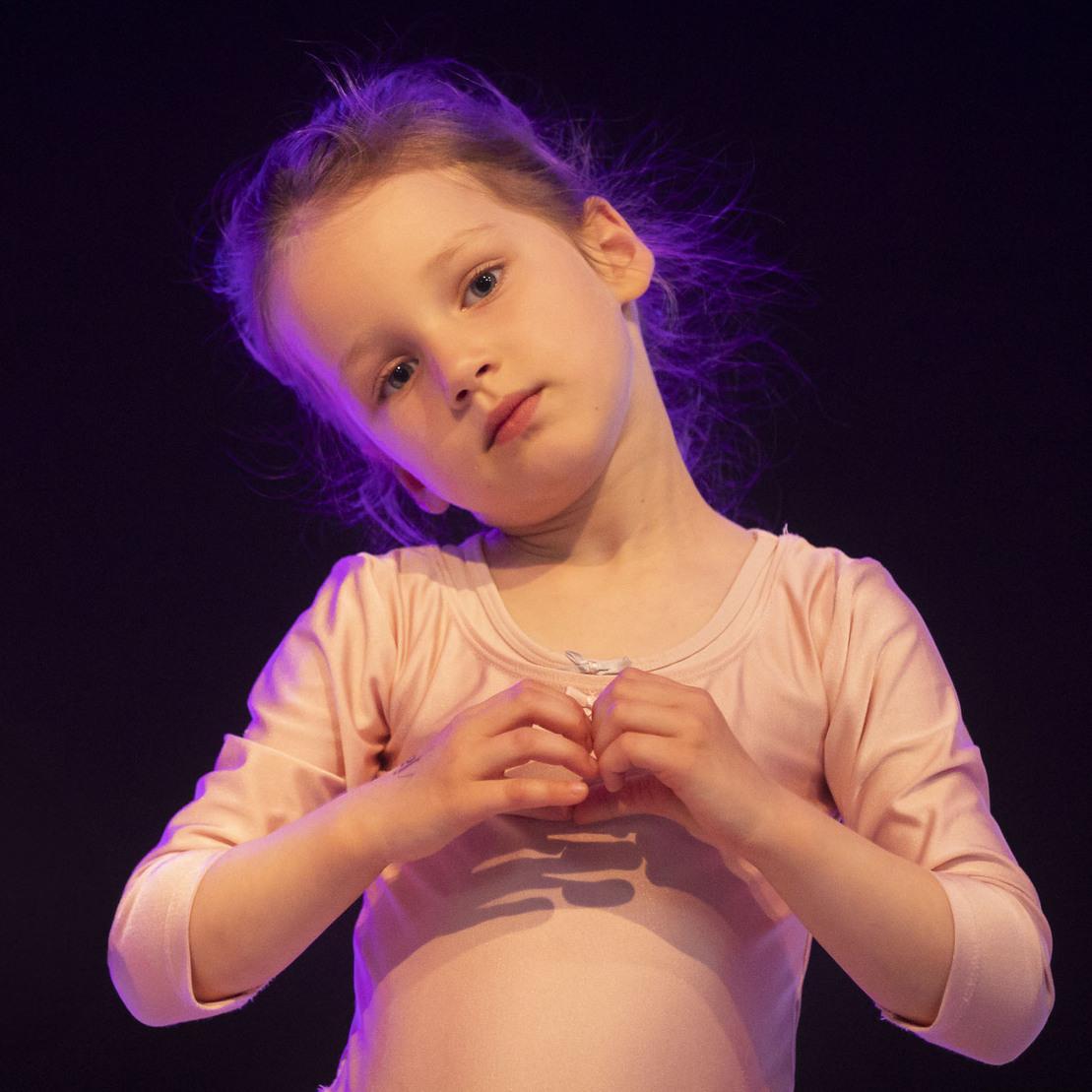 M&auml;dchen in rosa Ballettanzug formt mit H&auml;nden ein Herz vor dunklem Hintergrund.