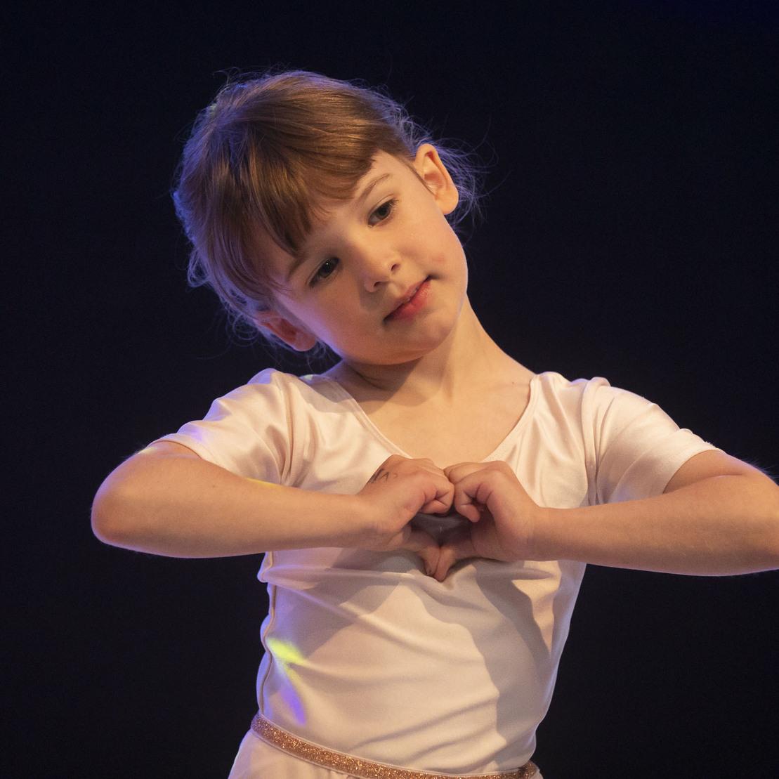 M&auml;dchen in Ballettkleidung formt mit ihren H&auml;nden ein Herz w&auml;hrend einer Auff&uuml;hrung auf dunkler B&uuml;hne.