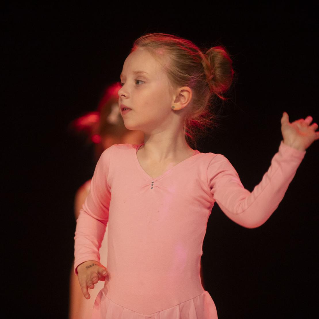 Tanzendes M&auml;dchen in rosa Ballettanzug, mit einer Hand erhoben, vor dunklem Hintergrund.