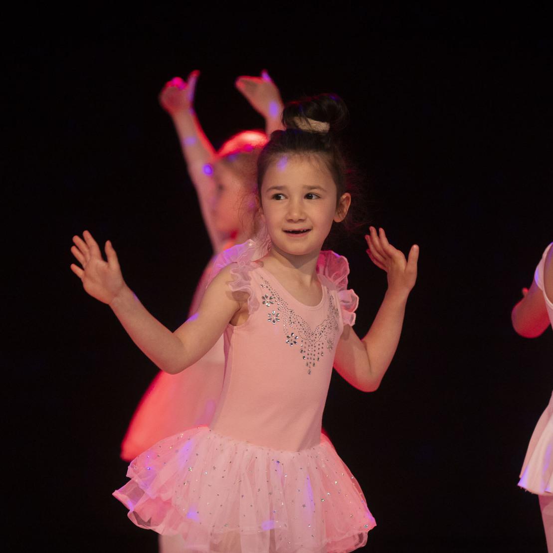 M&auml;dchen in rosa Ballettkost&uuml;m tanzt l&auml;chelnd auf einer B&uuml;hne mit bunten Lichtern.