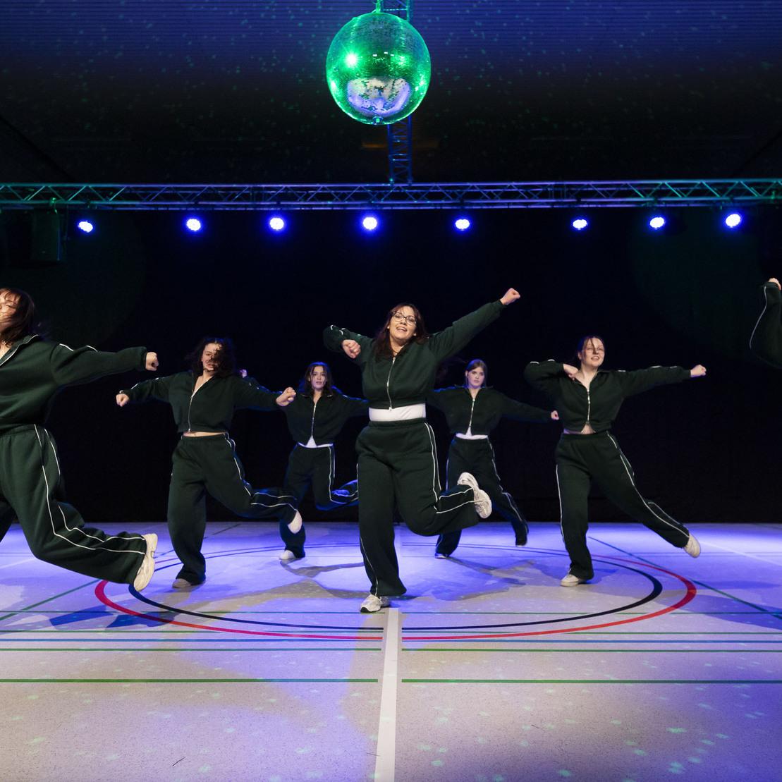 Tanzgruppe in dunkelgr&uuml;nen Outfits f&uuml;hrt eine Choreografie auf einer B&uuml;hne mit Disco-Kugel und bunten Lichtern auf.