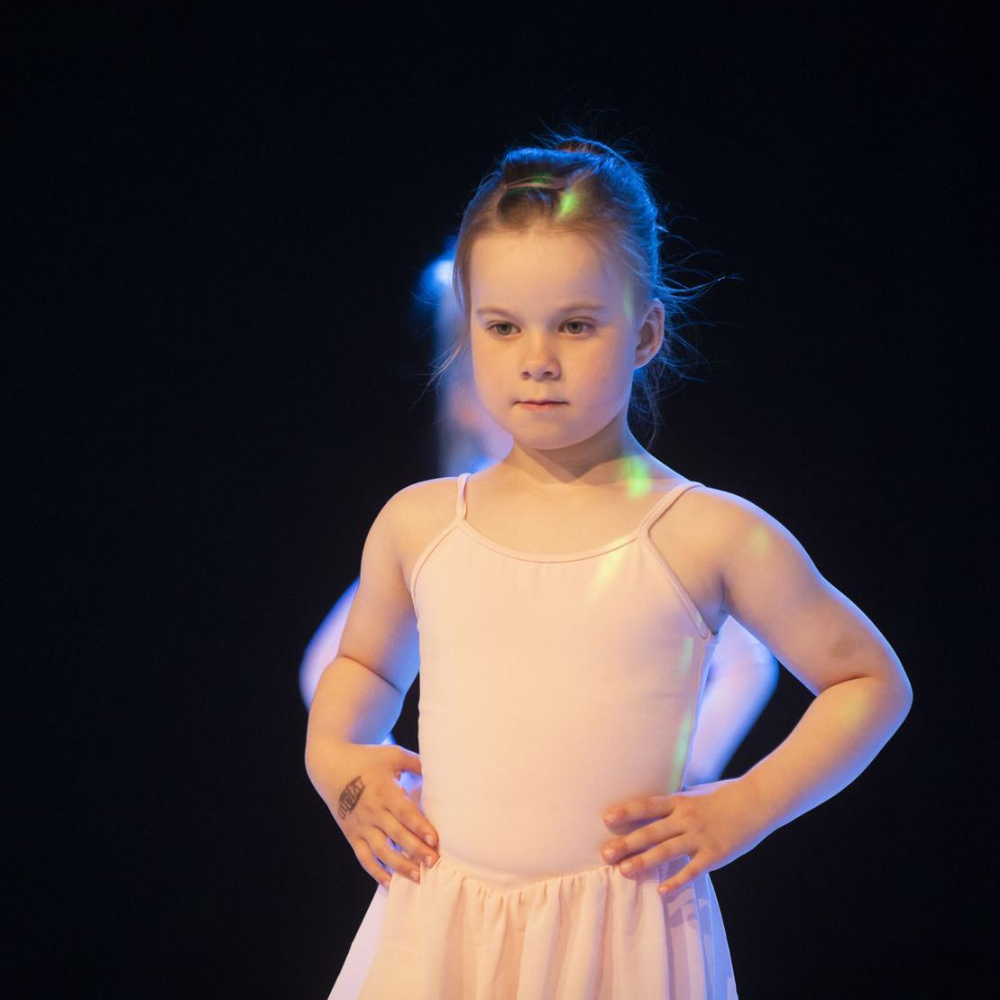 Junge Ballettt&auml;nzerin mit Zopf, die auf einer B&uuml;hne vor dunklem Hintergrund steht.