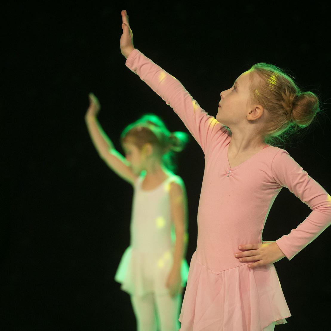 Zwei M&auml;dchen in Ballettkleidung tanzen auf einer B&uuml;hne, eines in Rosa, das andere in Wei&szlig;, mit angehobenem Arm.