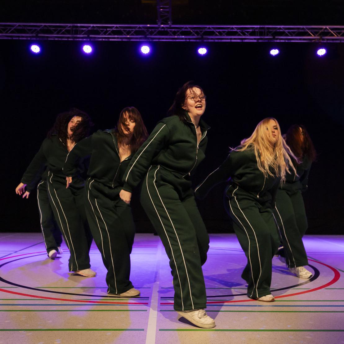 Sechs T&auml;nzerinnen in Sportanz&uuml;gen f&uuml;hren eine choreografierte Tanzbewegung auf einer beleuchteten B&uuml;hne aus.