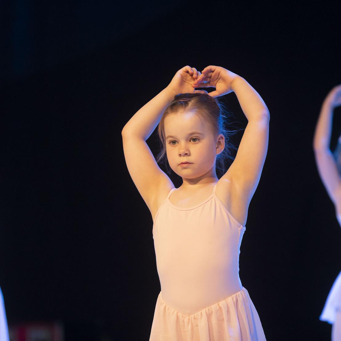 Drei kleine Ballett tanzende M&auml;dchen in rosa Tutus, eines mit erhobenen Armen, vor dunklem Hintergrund.