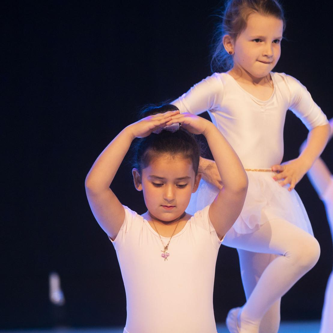 Drei T&auml;nzerinnen im wei&szlig;en Ballettanzkost&uuml;m pr&auml;sentieren eine Choreografie auf einer beleuchteten B&uuml;hne.