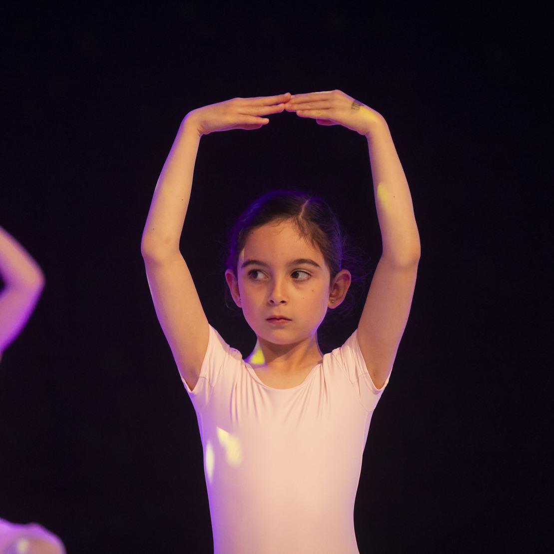 Tanzende M&auml;dchen in Ballettkleidung, eines hebt die Arme &uuml;ber den Kopf, der Hintergrund ist dunkel mit lila Lichtakzenten.
