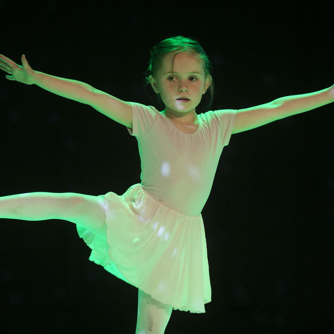 M&auml;dchen in einem wei&szlig;en Tanzkleid, das mit einem Bein in der Luft steht und die Arme weit ausbreitet, beleuchtet von gr&uuml;nem Licht.