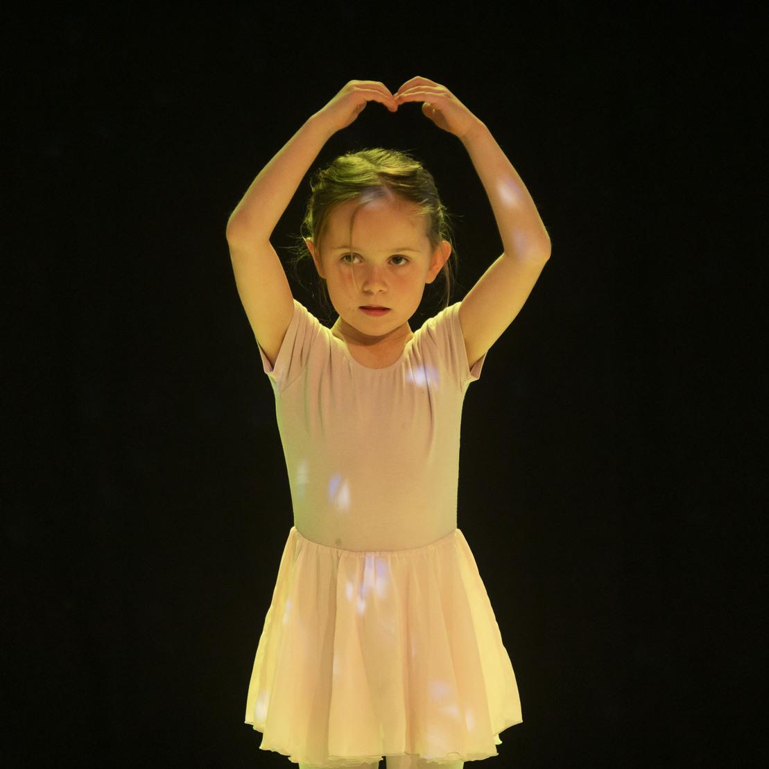 M&auml;dchen in Ballettkleidung formt mit den Armen ein Herz vor einem dunklen Hintergrund.