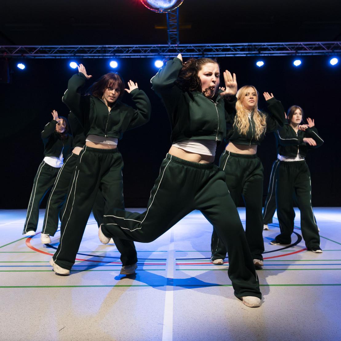 Tanzgruppe in gr&uuml;nen Outfits f&uuml;hrt eine energiegeladene Choreografie auf einer beleuchteten Fl&auml;che auf.